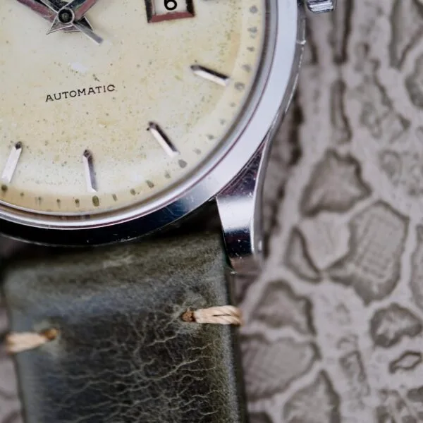 Close-up of an IWC Stainless Steel Automatic C.853 wristwatch featuring a cream face, round silver bezel, and black markers. The watch displays AUTOMATIC and a partial date function. It comes with a distressed green leather strap against a textured backdrop.