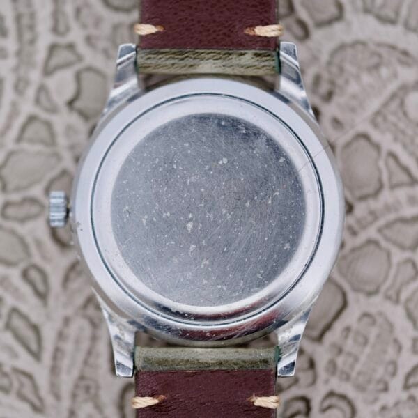 The image depicts the back of an IWC Stainless Steel Automatic C.853, a round silver wristwatch with a plain, scratched surface, attached to a brown leather strap featuring beige stitching, set against a textured background.