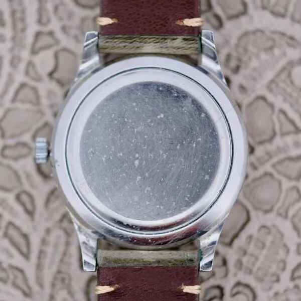 The image depicts the back of an IWC Stainless Steel Automatic C.853, a round silver wristwatch with a plain, scratched surface, attached to a brown leather strap featuring beige stitching, set against a textured background.