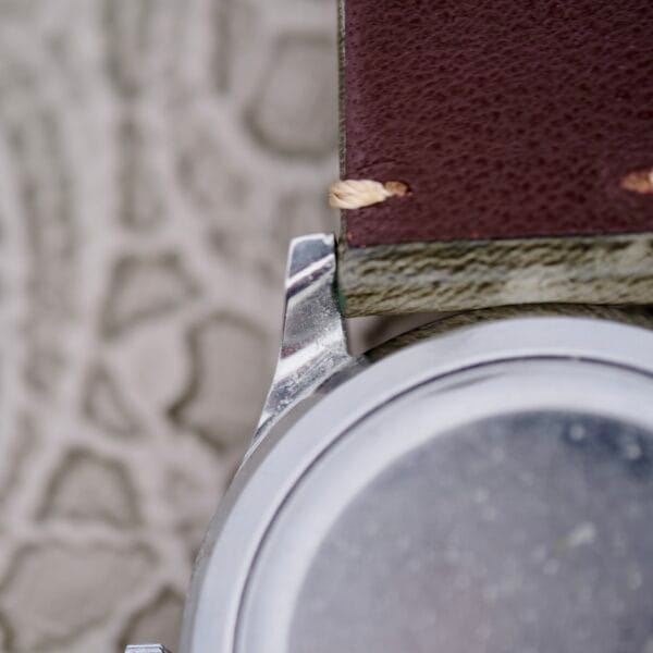 Close-up of the IWC Stainless Steel Automatic C.853 watch with a brown leather strap on a textured gray surface, highlighting the attachment area between the strap and watch body, showcasing stitching and metal details.