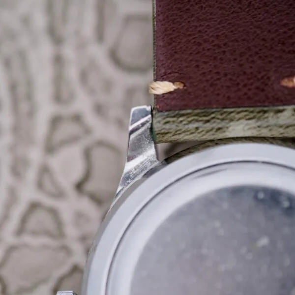 Close-up of the IWC Stainless Steel Automatic C.853 watch with a brown leather strap on a textured gray surface, highlighting the attachment area between the strap and watch body, showcasing stitching and metal details.