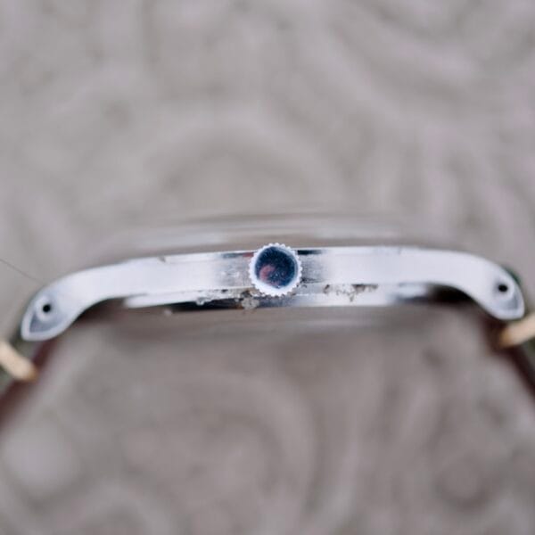 Close-up of an IWC Stainless Steel Automatic C.853s side profile shows the crown, a metallic finish with slight wear, two small screws on either side, and a leather strap. The blurred background features a textured pattern.