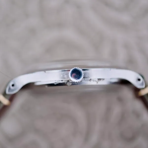 Close-up of an IWC Stainless Steel Automatic C.853s side profile shows the crown, a metallic finish with slight wear, two small screws on either side, and a leather strap. The blurred background features a textured pattern.