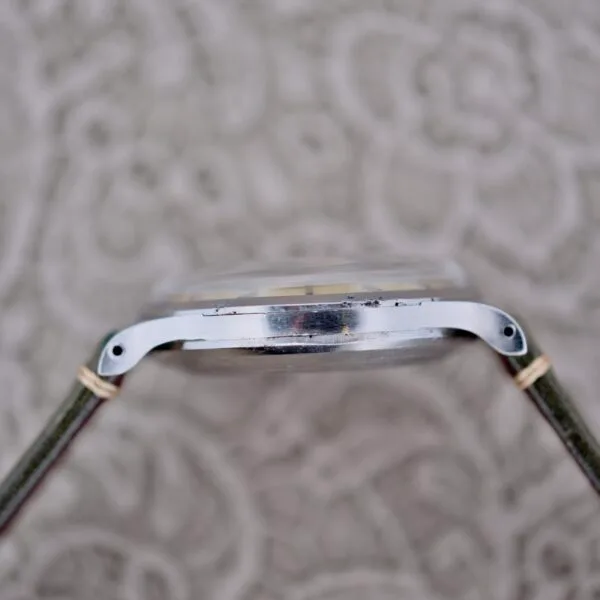 The IWC Stainless Steel Automatic C.853 is showcased in a side view against a patterned background, highlighting its thin metal case and domed crystal. The watchs green strap is partially visible as it rests at an angle.