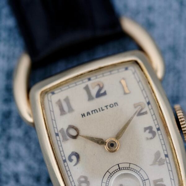 Close-up of a 1940s Hamilton Bowman wristwatch featuring a rectangular gold case with softly curved edges. It has a white face displaying Arabic numerals, hour and minute hands, and a small seconds dial, paired with a black leather strap.
