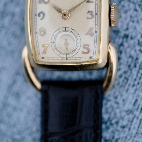 Close-up of a 1940s Hamilton Bowman vintage gold wristwatch with a rectangular face, black numerals, small seconds dial, and black leather strap, shown worn against grey textured fabric.