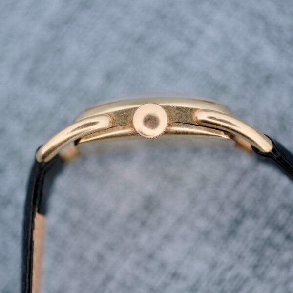 A side view of the 1940s Hamilton Bowman watch, featuring a gold case, round crown, and black strap, set against a textured gray background.