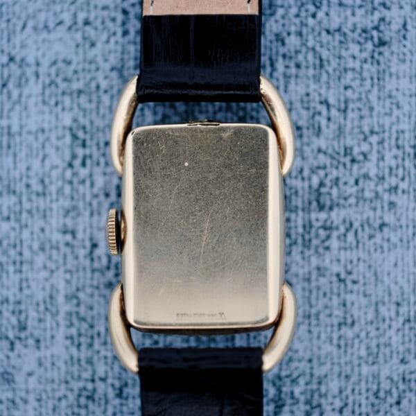 The 1940s Hamilton Bowman is a gold rectangular wristwatch with a black leather strap, shown from the back on a textured gray surface. It features a round winding crown and rounded gold lugs.
