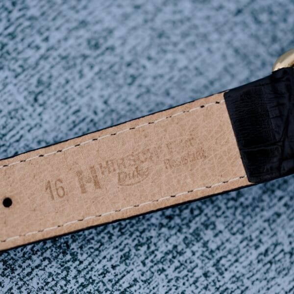 Close-up of a watch straps interior, showcasing tan leather imprinted with Hadley Roma Duke and 16. The strap has a black exterior and rests on a textured gray surface. Associated product: 1940s Hamilton Bowman.