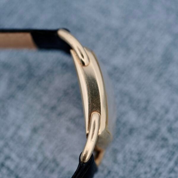 Close-up of a 1940s Hamilton Bowman gold watch with a black leather strap, showing the side view of its case against a textured gray surface.