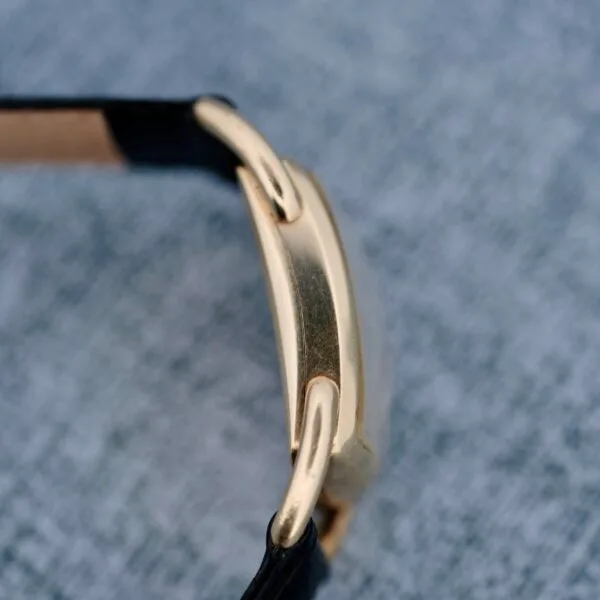 Close-up of a 1940s Hamilton Bowman gold watch with a black leather strap, showing the side view of its case against a textured gray surface.