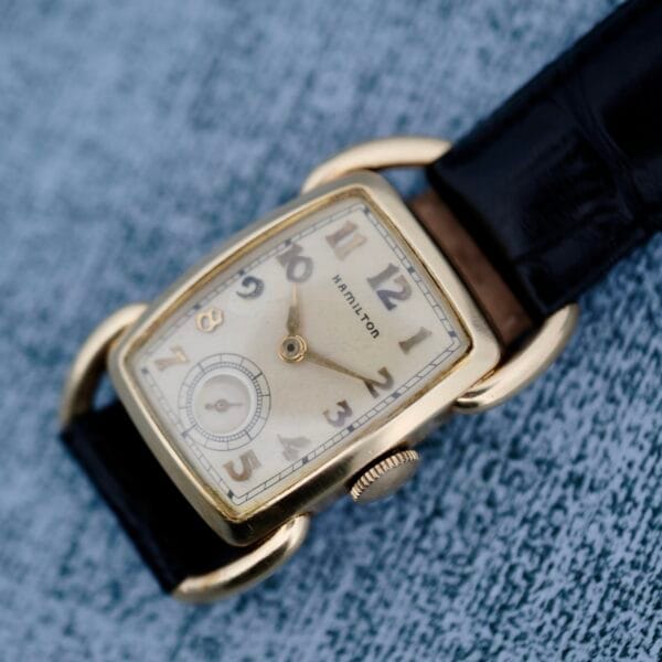 The 1940s Hamilton Bowman is a vintage wristwatch featuring a gold rectangular case and cream dial, gold Arabic numerals, a small seconds subdial at 6 oclock, and black leather straps, set against a textured gray surface.