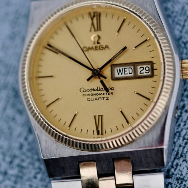 Close-up of an Omega Constellation Chronometer Quartz watch featuring a gold-toned face with Roman numerals at 12, 3, 6, and 9. The date window shows Wed 29, and the silver casing and two-tone bracelet have gold accents.