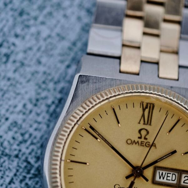 Close-up of an Omega Constellation Chronometer Quartz wristwatch showcasing a gold bezel and two-tone band. The face shows Roman numerals, the Omega logo, and a date window indicating WED and 2 against a textured blue background.