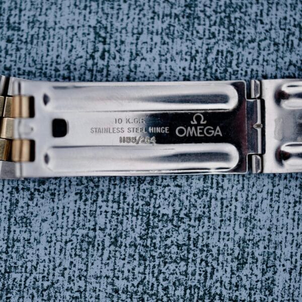 Close-up of a metal watch strap with engravings, featuring 10 K.G.F., STAINLESS STEEL HINGE, model number 0085/634, and the Omega brand. The Omega Constellation Chronometer Quartz watch has a combination of stainless steel and gold tones on the strap.