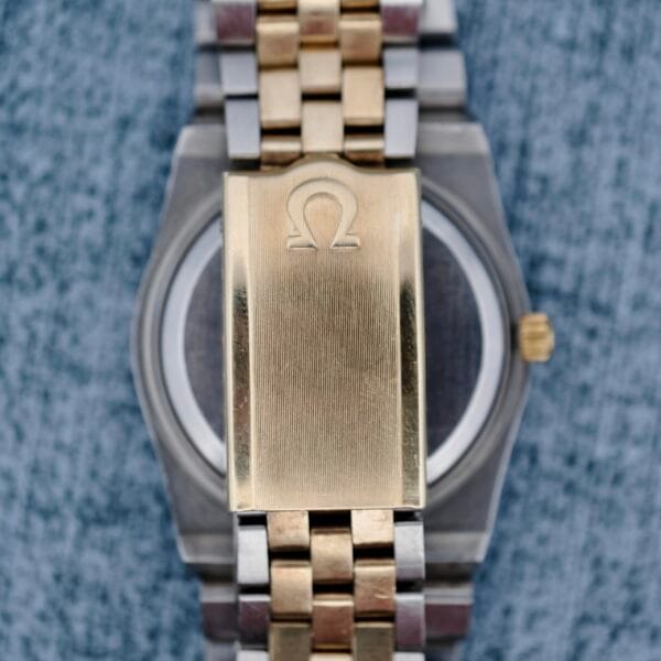 A close-up of the Omega Constellation Chronometer Quartz bracelet shows two-tone metal links in gold and silver, with a gold clasp embossed with the Omega logo against a textured bluish-gray background.