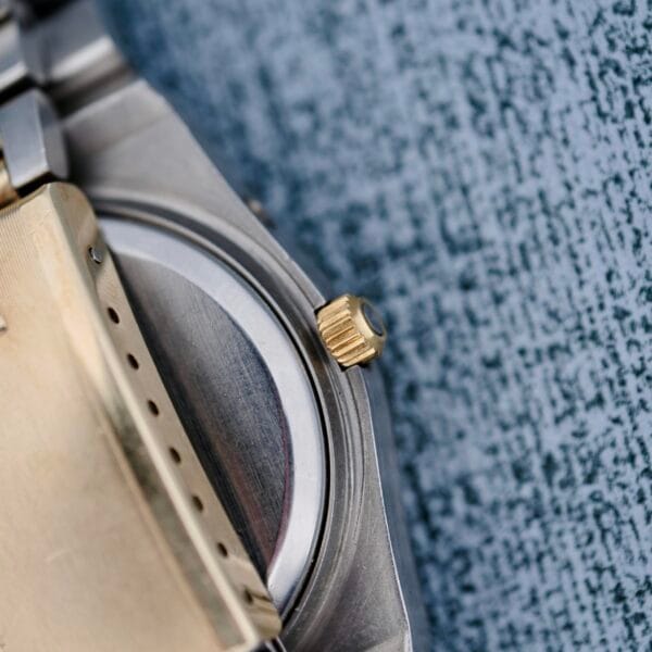 Close-up of the Omega Constellation Chronometer Quartz underside, featuring a gold crown and part of its stainless steel and gold case against a textured blue-gray background. The metal bracelet is visible on the left.