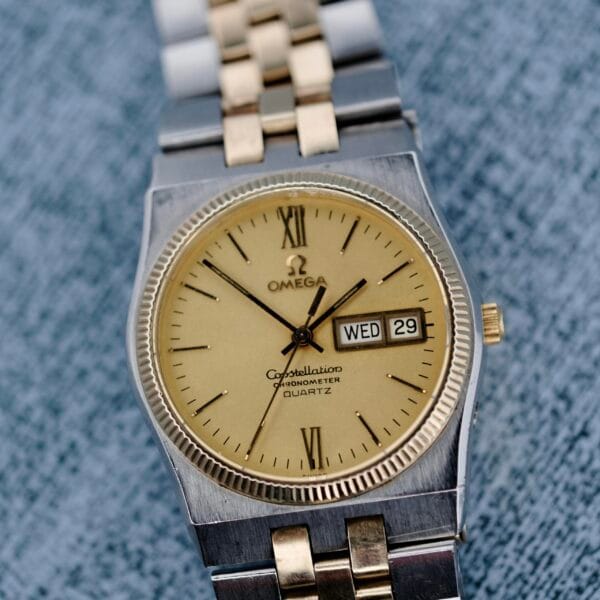 Close-up of an Omega Constellation Chronometer Quartz watch featuring a stainless steel and gold-tone bracelet. It has a beige dial with Roman numerals, a day/date window showing Wed 29, ridged bezel, and textured blue background.