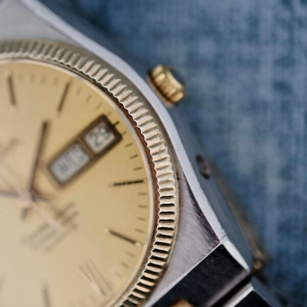 Close-up of an Omega Constellation Chronometer Quartz wristwatch, featuring a gold textured bezel and champagne dial with day and date set to Wednesday the 28th. The background is a blurred blue-gray surface.