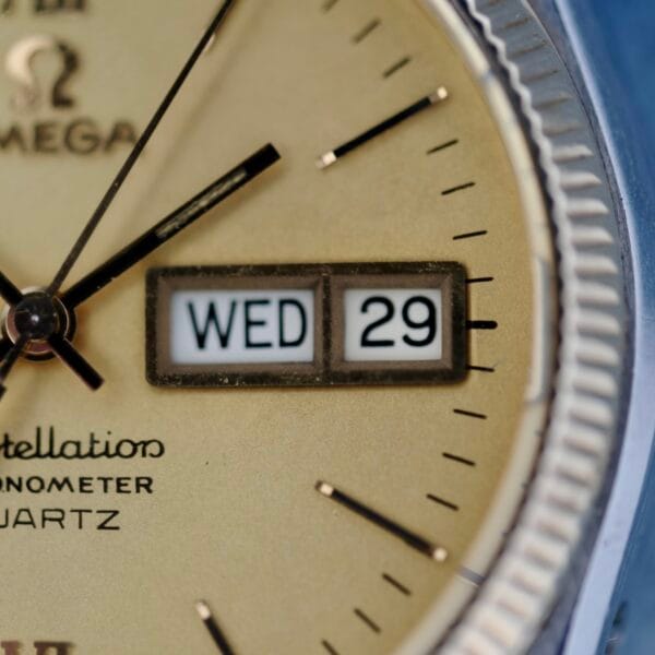 Close-up of a vintage Omega Constellation Chronometer Quartz watch face, showing approximately 7:48. A beige background features black hour markers. The day-date window shows WED 29, and the Omega logo with text is clear on the dial.