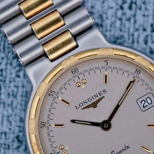 Close-up of a Longines Conquest Quartz Two-Tone 4940 watch on a textured surface, featuring a silver and gold link bracelet, cream dial, gold-tone markers, date window displaying the 20th, black hands, and logo beneath the brand name.