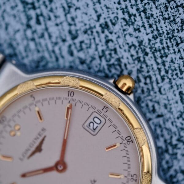 Close-up of the Longines Conquest Quartz Two-Tone 4940 wristwatch with a gold and silver bezel on a textured blue surface, featuring a date window set to the 20th and gold hour markers and hands.