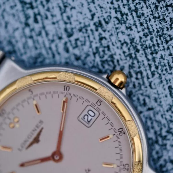 Close-up of the Longines Conquest Quartz Two-Tone 4940 wristwatch with a gold and silver bezel on a textured blue surface, featuring a date window set to the 20th and gold hour markers and hands.