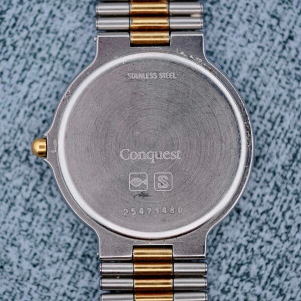 The image shows the back of a Longines Conquest Quartz Two-Tone 4940 stainless steel watch with a metal band, engraved with STAINLESS STEEL, Conquest, two symbols, and the number 25971480. The surface appears slightly scratched and worn.
