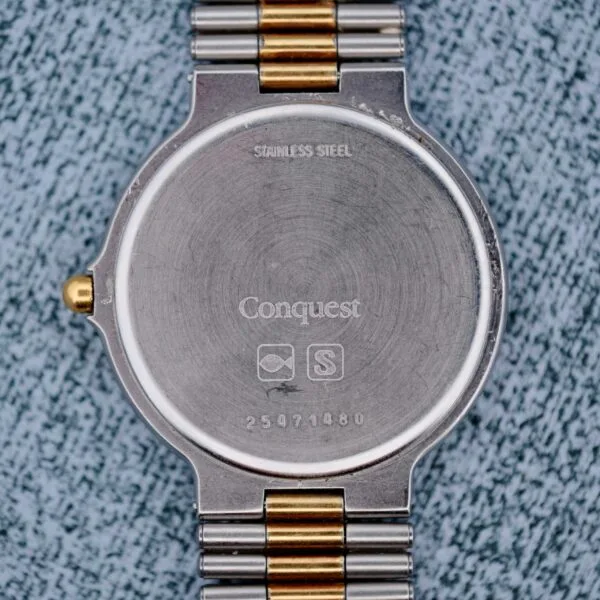 The image shows the back of a Longines Conquest Quartz Two-Tone 4940 stainless steel watch with a metal band, engraved with STAINLESS STEEL, Conquest, two symbols, and the number 25971480. The surface appears slightly scratched and worn.
