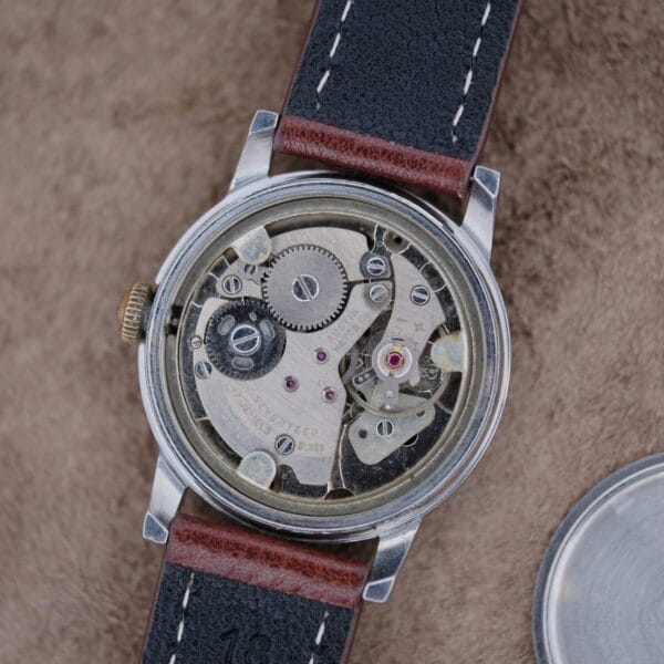 A close-up of the Austin Deluxe 17 Jewels Blue Patina Dial wristwatch with its back cover removed reveals exposed gears. The watch features a silver case, brown and black leather strap, and rests on a textured brown surface.