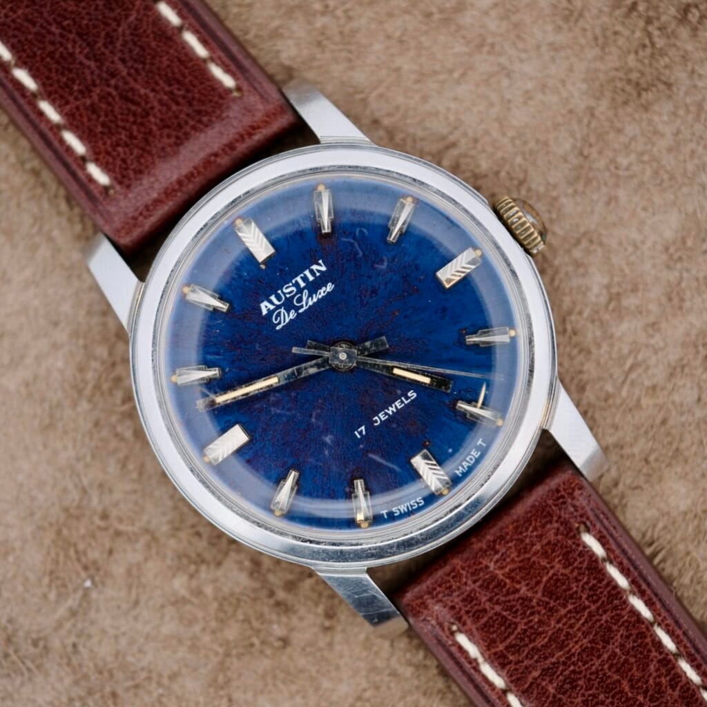 The Austin Deluxe 17 Jewels Blue Patina Dial wristwatch, featuring a brown leather strap, is elegantly displayed on a beige surface.