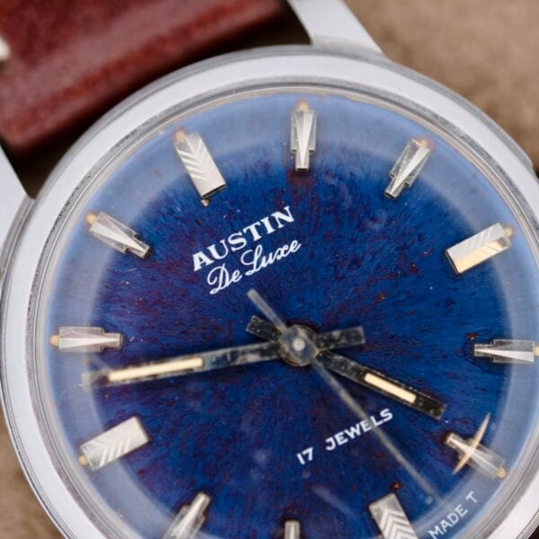 Close-up of the Austin Deluxe 17 Jewels Blue Patina Dial wristwatch, featuring silver hour markers and hands on a textured blue face, plus a partially visible brown leather strap.
