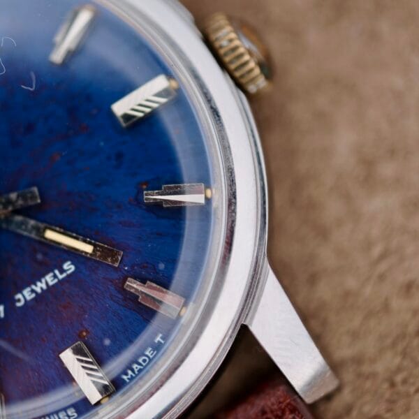 Close-up of the Austin Deluxe 17 Jewels Blue Patina Dial wristwatch, featuring a blue dial with silver hour markers, part of a gold crown, a silver case, and a brown leather strap. JEWELS and MADE T are visible on the dial.