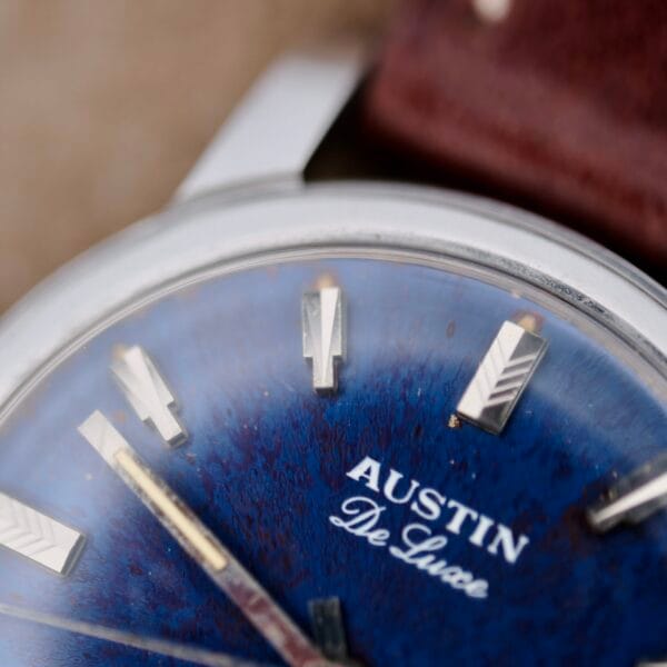 A close-up of the Austin Deluxe 17 Jewels Blue Patina Dial watch shows a dark blue textured face with silver hour markers and AUSTIN De Luca on the dial, along with a glimpse of its brown leather strap.