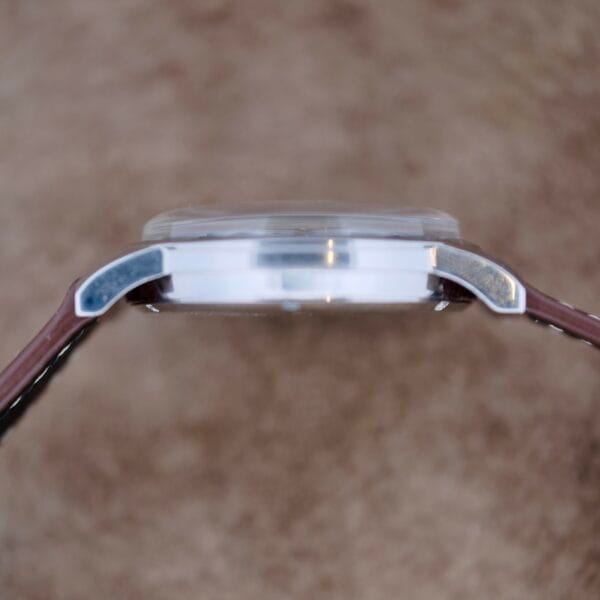 A close-up side view of the Austin Deluxe 17 Jewels Blue Patina Dial wristwatch shows its clear, slightly domed crystal and brown leather strap, set against a blurred neutral background.