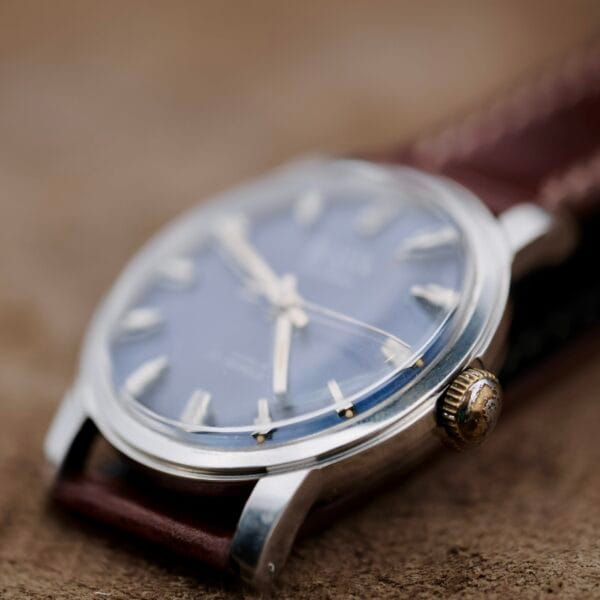 A close-up of the Austin Deluxe 17 Jewels with a silver case, brown leather strap, and blue patina dial, gold markers in focus, resting on a textured brown surface.