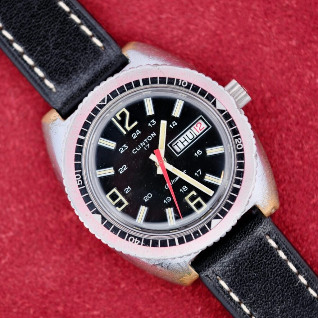 A Clinton World Timer Automatic Day/Date watch with a black leather strap displayed on a red textured background.