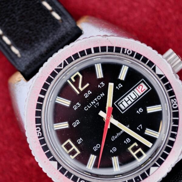 A close-up of the Clinton World Timer Automatic Day/Date watch with a black face, white and gold hour markers, red second hand, black leather strap, showing Thursday the 12th. The background is red.