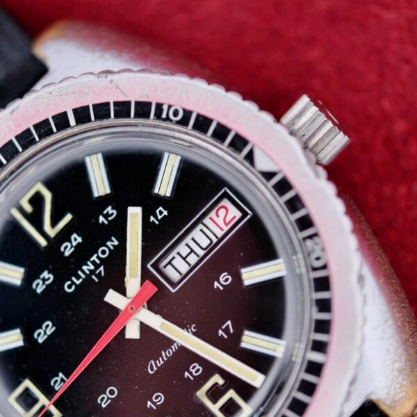 Close-up of a Clinton World Timer Automatic Day/Date watch displaying THU 12. It features a black face, silver bezel, white and yellow hour markers, red second hand, and is set against a red background.
