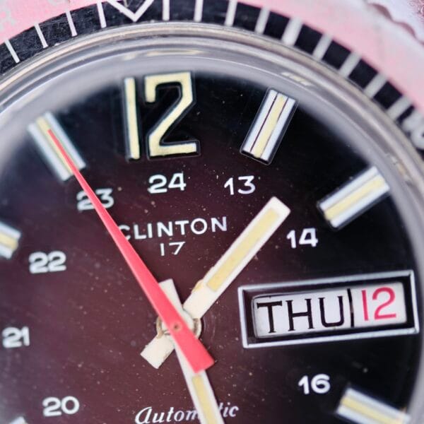 Close-up of a Clinton World Timer Automatic Day/Date watch showing 11:57, with both 12-hour and 24-hour markings, the brand CLINTON, and a date window displaying THU 12.