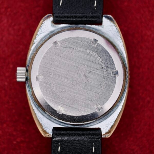 The back of the Clinton World Timer Automatic Day/Date stainless steel watch with a black leather strap is shown against a red background, featuring an engraved caseback with scratches, left-positioned crown, and partially visible lugs.