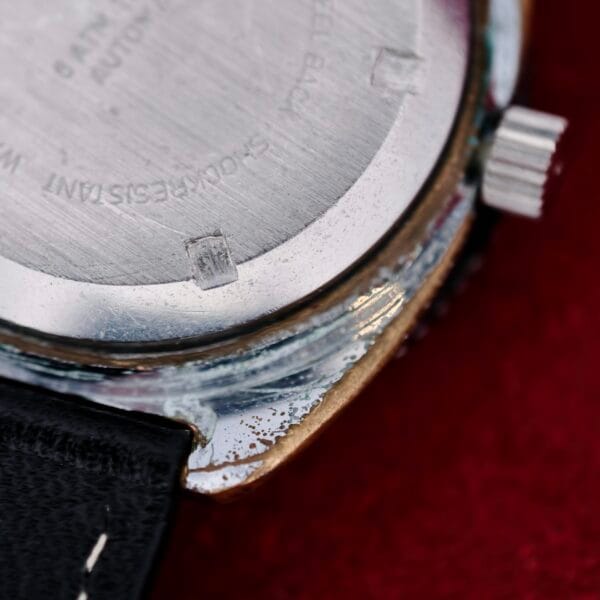 Close-up of the back of a Clinton World Timer Automatic Day/Date, showing scratches and wear, with part of the black leather strap and crown visible against a red background.