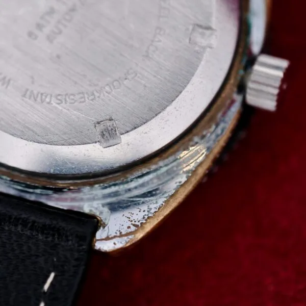 Close-up of the back of a Clinton World Timer Automatic Day/Date, showing scratches and wear, with part of the black leather strap and crown visible against a red background.
