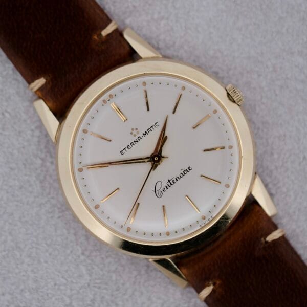 A 1950s Eterna Centaire Eterna Matic Cal. 1428U wristwatch with a gold case, white dial, gold hour markers and hands, and a brown leather strap is displayed on a light gray surface.