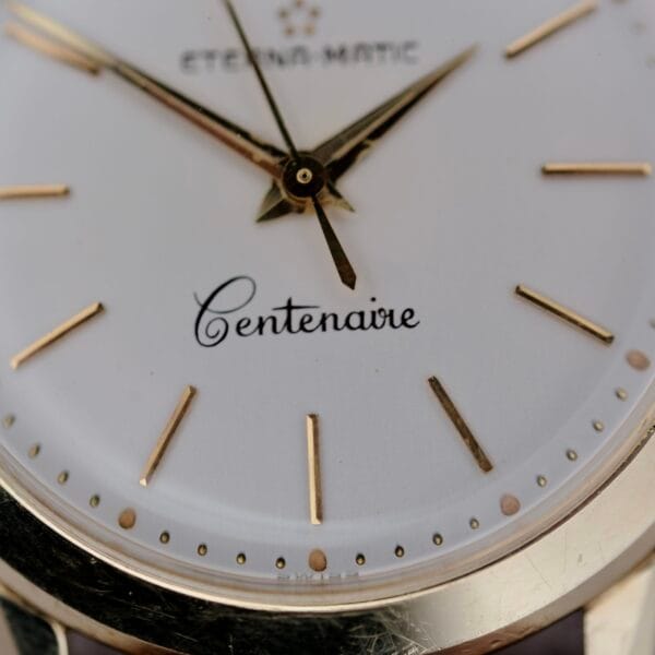 Close-up of a 1950s Eterna Centenaire Eterna-Matic Cal. 1428U wristwatch face featuring the brand name at the top, Centenaire in cursive at the center, and gold hour markers and hands on a white background.