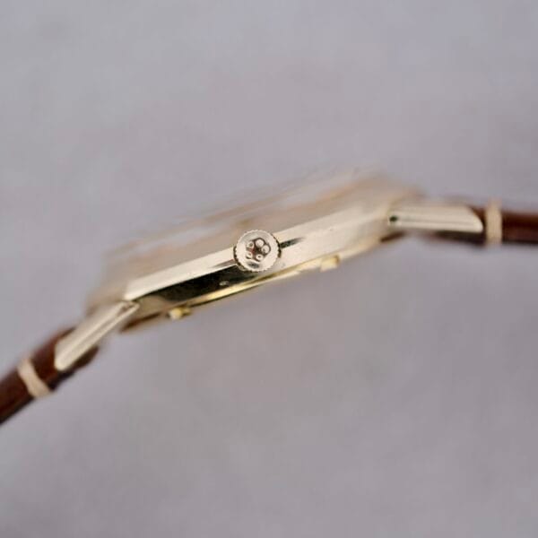 A close-up side view of the 1950s Eterna Centaire Eterna Matic Cal. 1428U gold wristwatch with a brown leather strap, focusing on the crown against a plain, light gray background.