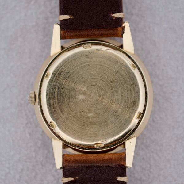 A 1950s Eterna Centaire Eterna Matic Cal. 1428U with a gold-colored round case and brown leather strap is displayed from the back on a light gray textured surface, showing no markings or engravings.