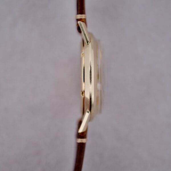 A close-up side view of a 1950s Eterna Centaire Eterna Matic Cal. 1428U gold wristwatch case with a brown leather strap, highlighting its thin profile against a plain light gray background.