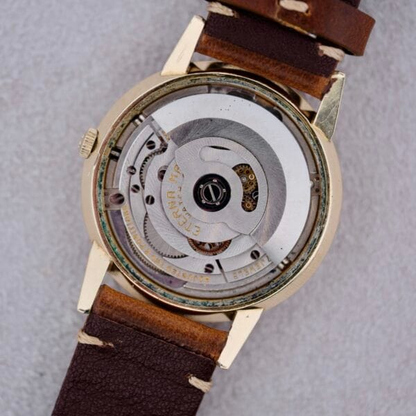 Close-up of the back of a 1950s Eterna Centaire Eterna Matic Cal. 1428U wristwatch, featuring an exposed mechanical movement, brown leather strap, gold-hued case, intricate gears, and engraved detailing.