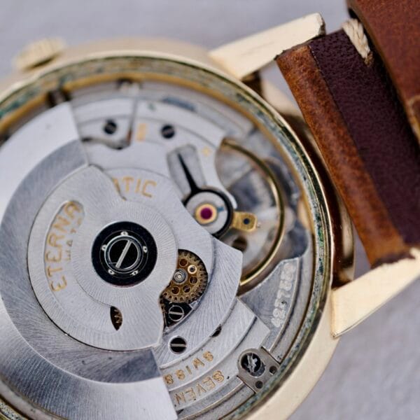 Close-up view of the 1950s Eterna Centaire Eterna Matic Cal. 1428U’s inner mechanical movement, showcasing visible gears and components, alongside the edge of a brown leather strap and part of its gold-toned case.